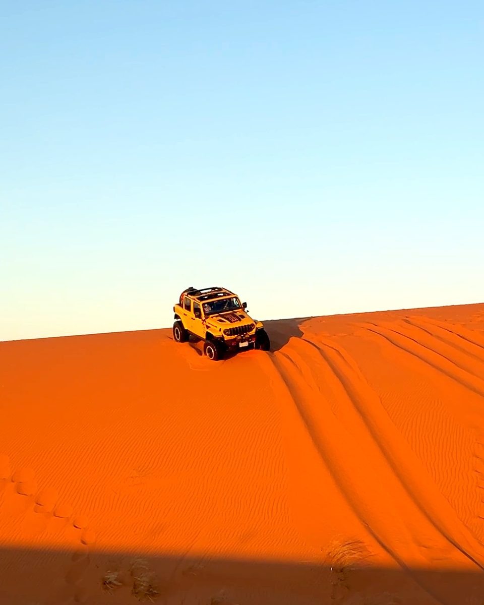 A yellow 4x4 on the crest of a red dune against a clear sky